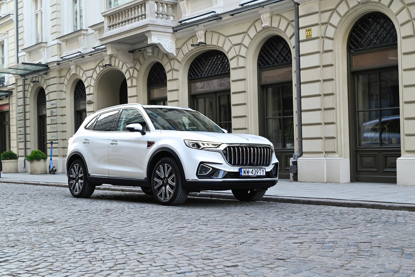 Biały nowoczesny SUV zaparkowany na brukowanej ulicy przed elegancką kamienicą z klasyczną architekturą. W tle widoczne duże okna i zdobione elementy fasady.