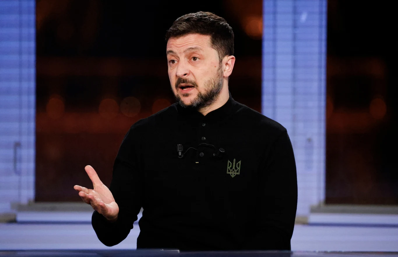 Zełenski grzmi po ataku Rosji na Ukrainę Mężczyzna w czarnym ubraniu siedzi przy stole, wykonując gest dłonią. Za nim widać okno z jasno oświetlonym, ale niewyraźnym tłem. Na jego ubraniu znajduje się logo z literami 'UA'. Wygląda na osobę zaangażowaną w rozmowę lub dyskusję.