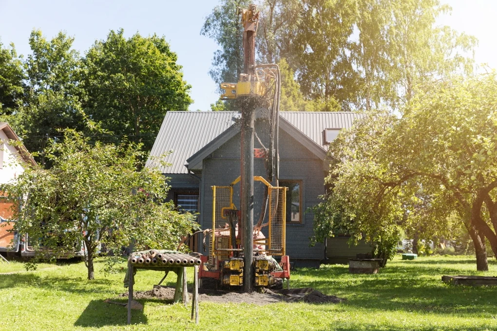 Coraz więcej osób budujących dom lub planujących zakup działki rekreacyjnej rozważa inwestycję we własne ujęcie wody Wiercenie geotermalnego odwiertu na terenie przydomowym. Na pierwszym planie widać wiertnicę, dalej dom mieszkalny, otoczony zielenią i drzewami. Jasne, słoneczne światło, które dodaje scenerii pogodnego charakteru.