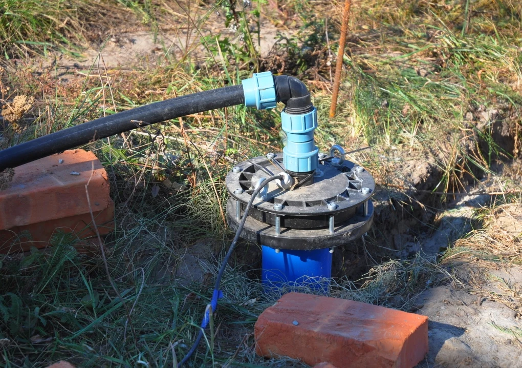 Jak wybrać odpowiednią pompę? Instalacja wodna na zewnątrz z widocznymi plastikowymi rurami i przyłączem, otoczona cegłami i roślinnością.