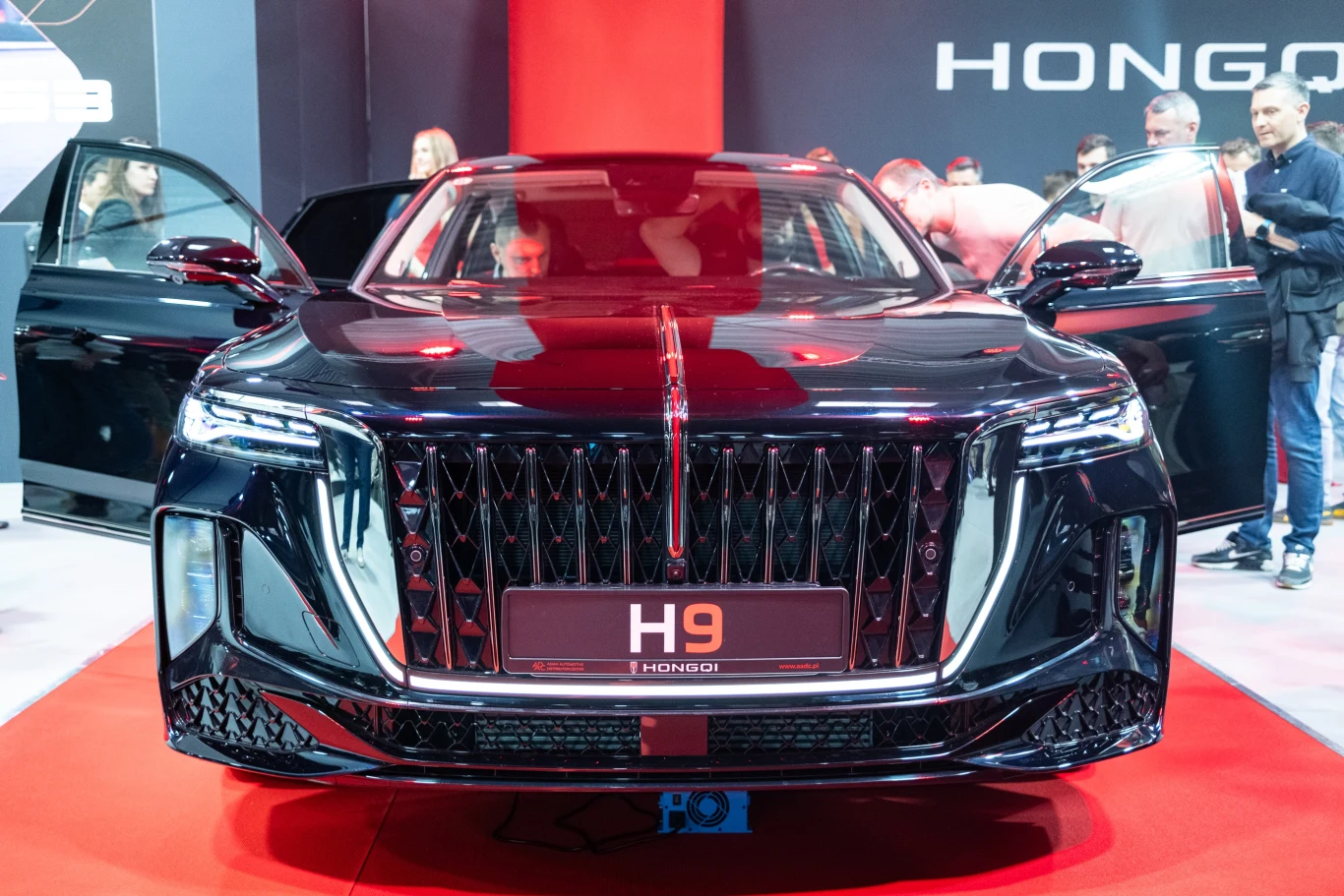 Luksusowy samochód marki Hongqi H9 prezentowany na targach motoryzacyjnych. Wyróżnia się elegancką, czarną karoserią z charakterystycznym grillem. W tle widoczni są ludzie przyglądający się pojazdowi.