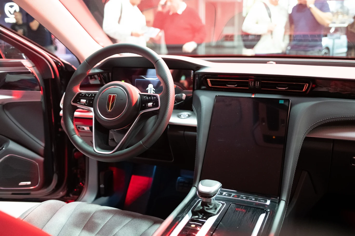 Wnętrze nowoczesnego samochodu z widocznym kokpitem, dużym ekranem dotykowym na konsoli środkowej i elegancką kierownicą. Czerwone refleksje podkreślają nowoczesność i styl pojazdu.