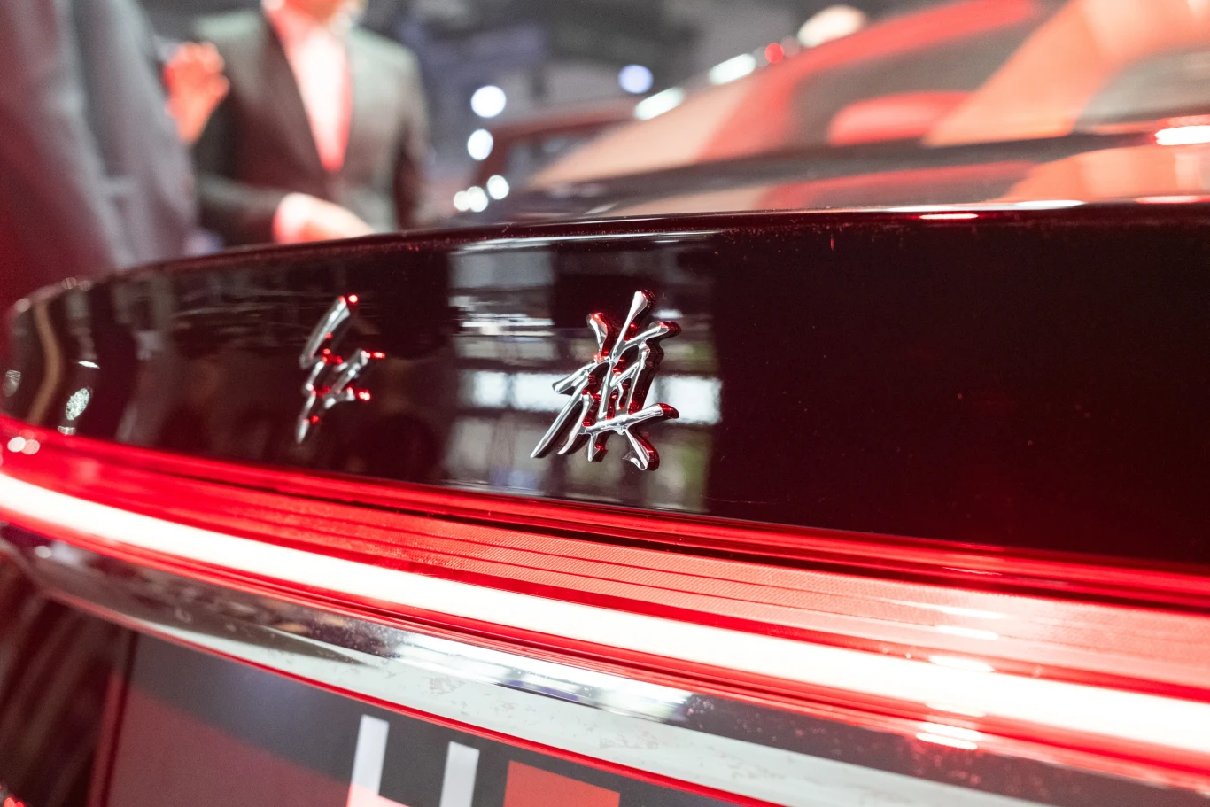 Czerwony samochód ze srebrnym oznaczeniem na tyle, z widocznymi detalami i rozmytą postacią w tle. Elegancki design i nowoczesne oświetlenie wskazują na luksusowy charakter pojazdu.