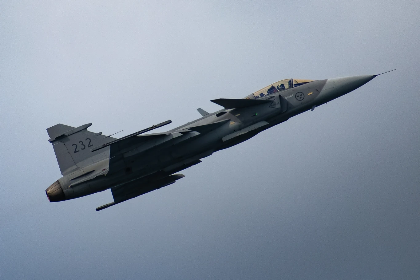 Myśliwiec JAS-39C Gripen Wojskowy samolot myśliwski w locie, widoczny pod kątem, ukazujący detale konstrukcji i oznaczeń na kadłubie, na tle nieba.