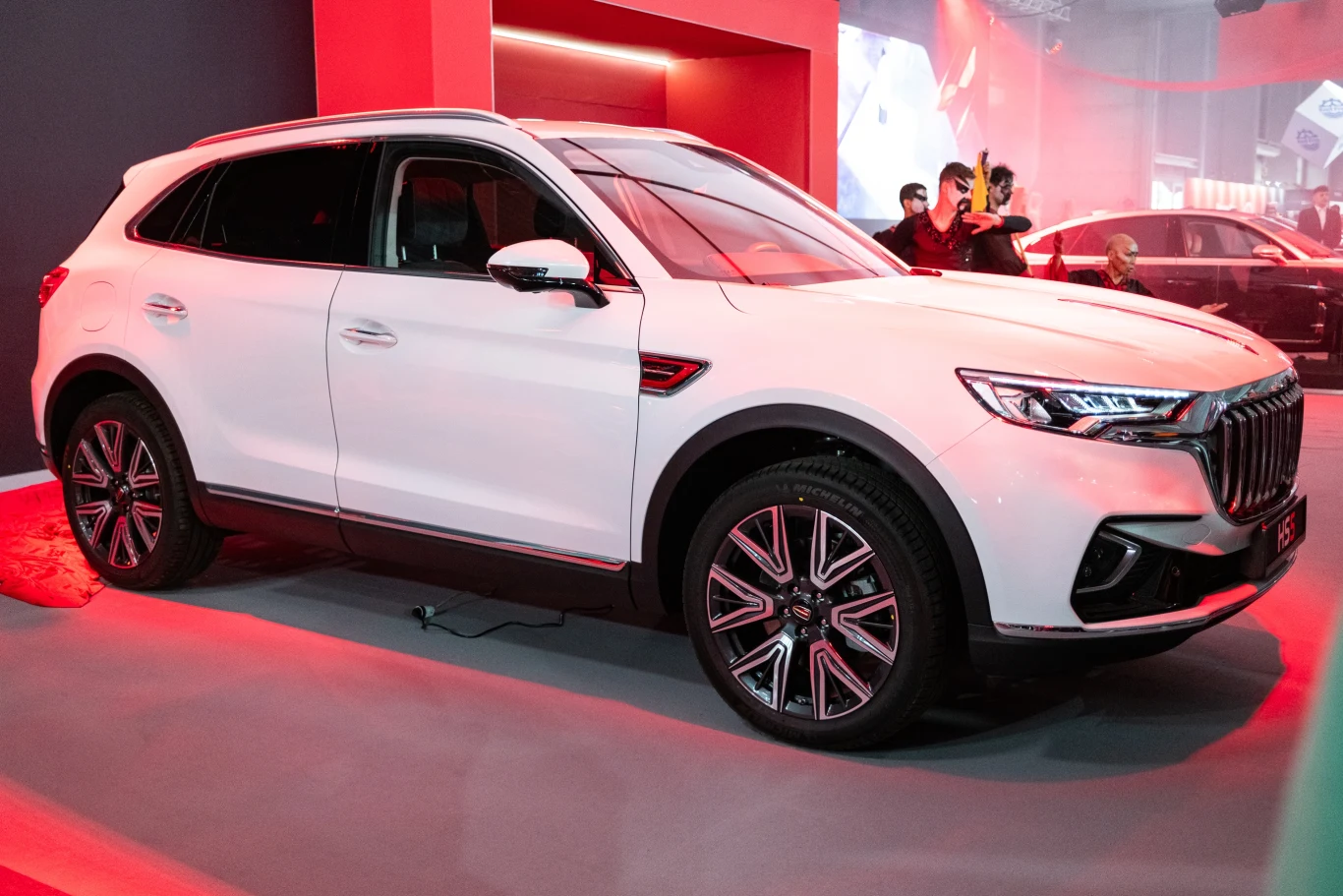 Białe, nowoczesne SUV stoi wystawione na targach motoryzacyjnych. Samochód ma elegancki design z wyraźnie oznaczonymi reflektorami i chromowanymi detalami. Tło stanowi jasne, rozświetlone stoisko z innymi autami w tle.