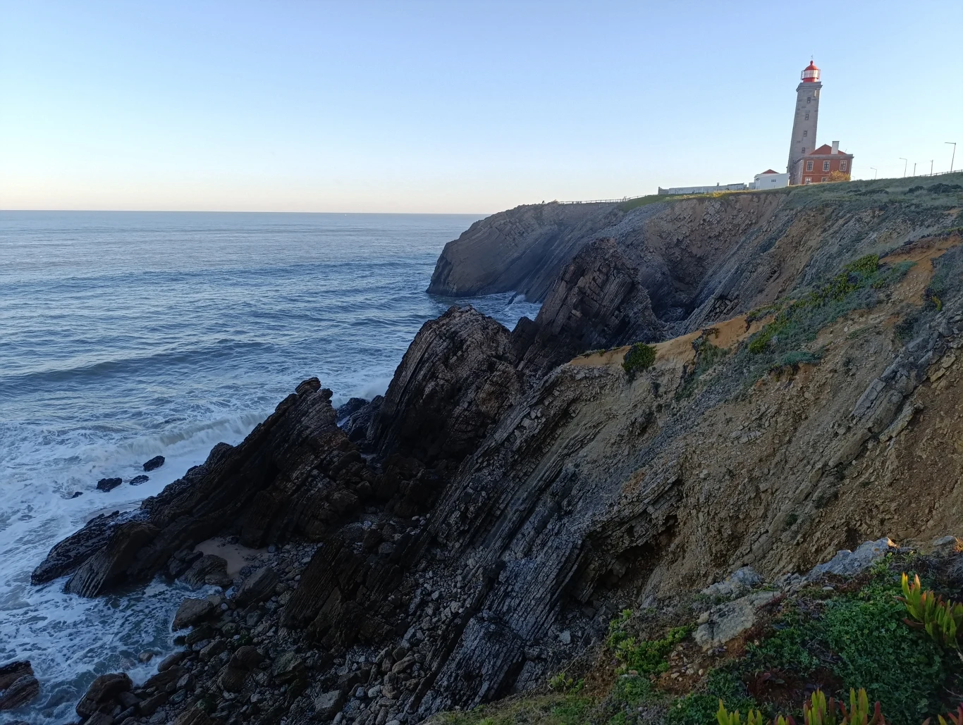 Latarnia morska na klifie nad brzegiem morza, z falami uderzającymi o skaliste wybrzeże pod niebem w odcieniach błękitu.
