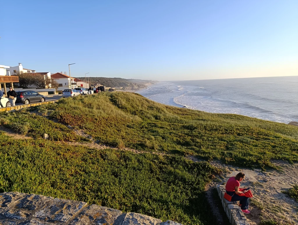 Takie widoki możecie mieć codziennie w São Pedro de Moel Szeroka panorama nadmorskiego krajobrazu z oceanem po prawej stronie, zalesioną skarpą pośrodku i osiedlem z domami po lewej. Na przodzie po prawej osoba siedzi w trawie, prawdopodobnie czytając.