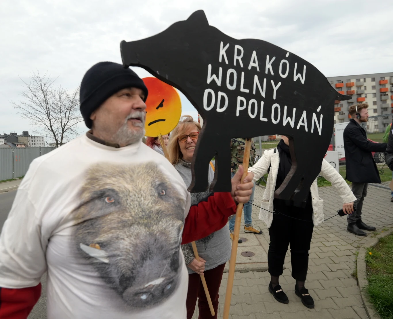 Grupa osób podczas protestu, mężczyzna trzymający czarny tekturowy znak w kształcie dzika z napisem 'Kraków wolny od polowań'. Osoba ubrana w koszulkę z wizerunkiem dzika.