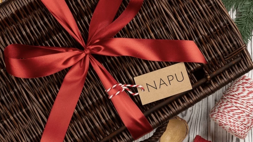 napu.pl oferuje ekspresową dostawę nawet w 24 godziny Kosz wiklinowy ozdobiony czerwoną, satynową wstążką i bilecikiem z napisem 'NAPU', umieszczony na jasnym tle drewnianym. Obok widoczne są sznurek w czerwono-białym kolorze i fragment zielonej gałązki.