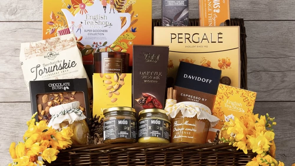 napu.pl Kosz delikatesowy z różnymi smakołykami i napojami, umieszczony na tle ozdób kwiatowych. W koszu znajdują się produkty takie jak herbata, czekolada, kawa, słoiki z miodem oraz orzechami w czekoladzie.