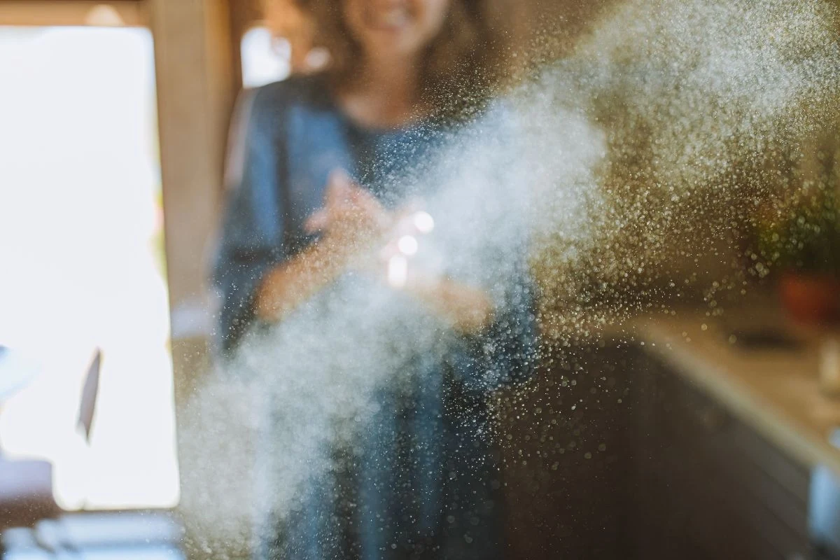 Rozproszony pyłek lub mąka w powietrzu w domowym wnętrzu, z unoszącymi się drobinkami w promieniach światła. W tle widoczna niewyraźna sylwetka kobiety.