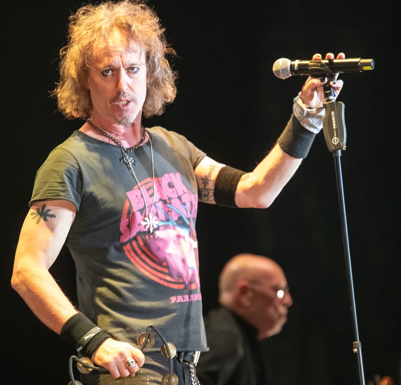 Carl Sentance (Nazareth) w akcji Mężczyzna na scenie trzyma mikrofon w jednej ręce, drugą ręką trzyma tamburyn. Ubrany jest w koszulkę z grafiką i biżuterię. W tle, drugi mężczyzna widoczny z profilu.