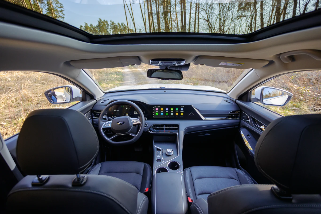 Wnętrze nowoczesnego samochodu z panoramicznym dachem, widok na deskę rozdzielczą z dużym ekranem dotykowym. Siedzenia pokryte czarną skórą, przestronne i komfortowe. Na zewnątrz widać las i jasne niebo.