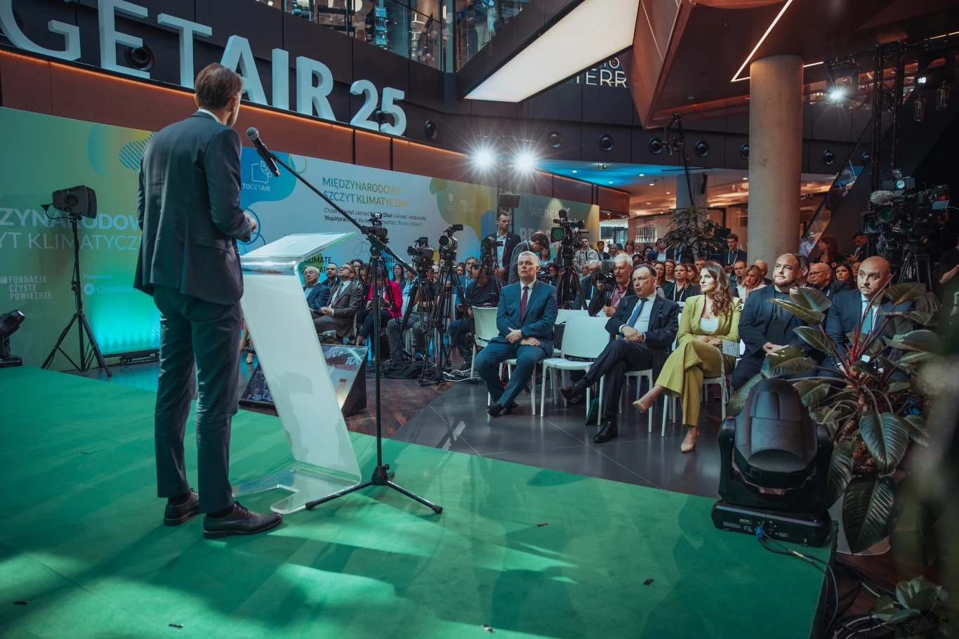 Szczyt TOGETAIR 2025 Przemówienie na konferencji, przy mównicy stoi mężczyzna, za nim widownia siedząca na krzesłach, ustawione kamery i urządzenia rejestrujące, w tle baner z napisem 'Get Air 25'.