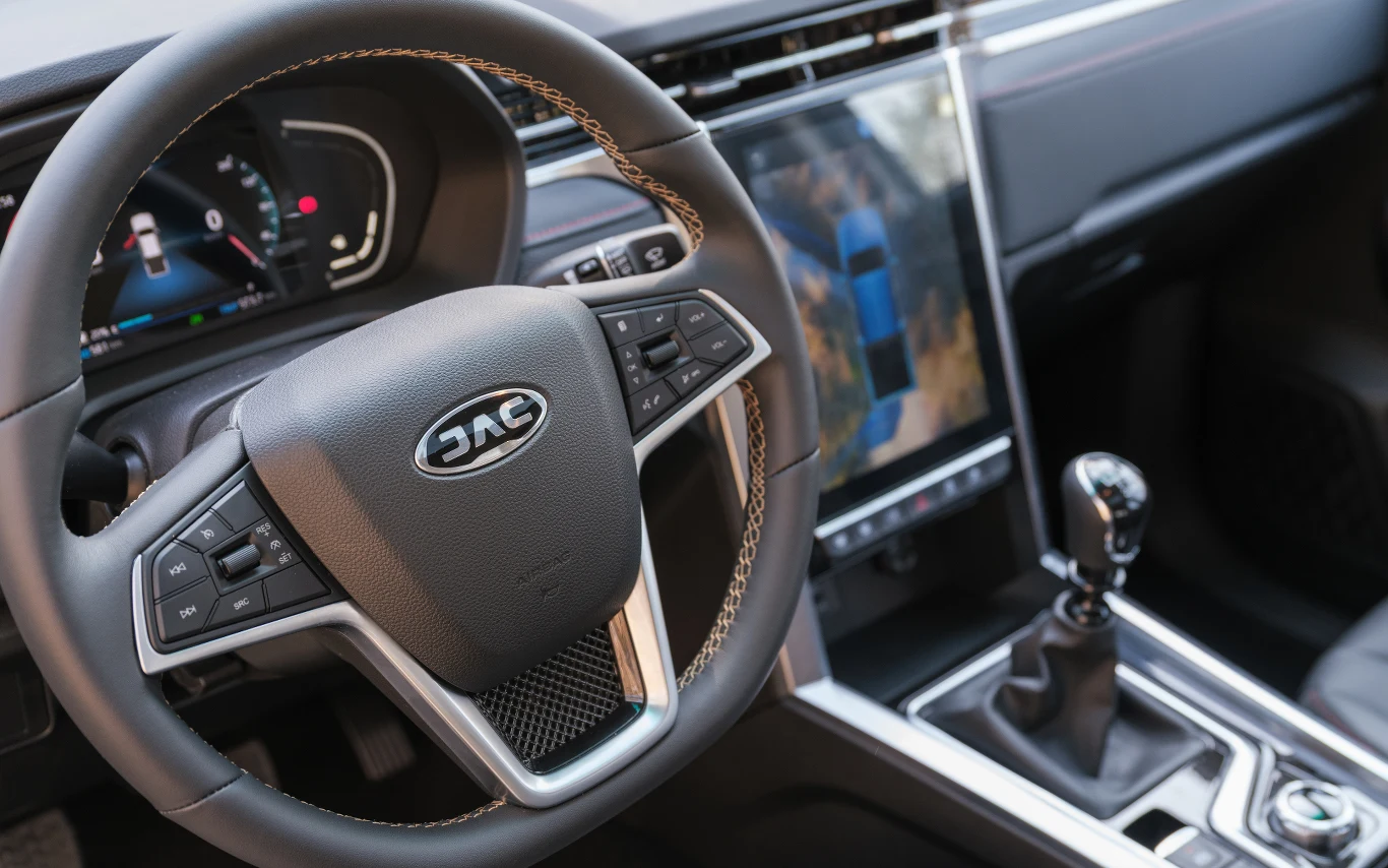 Wnętrze samochodu z widocznym kierownicą marki JAC, nowoczesną deską rozdzielczą z cyfrowym wyświetlaczem oraz dźwignią zmiany biegów.