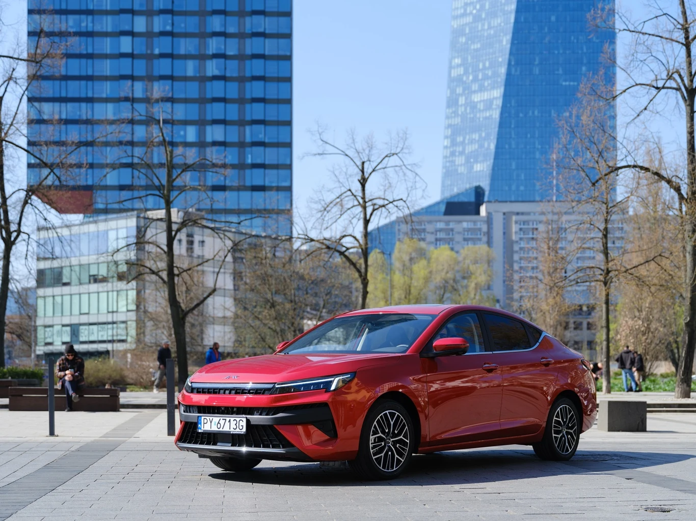 Czerwony sedan zaparkowany na tle nowoczesnych wieżowców w przestrzeni miejskiej. Słoneczna pogoda podkreśla eleganckie linie samochodu. W tle widać ludzi spacerujących po parku.