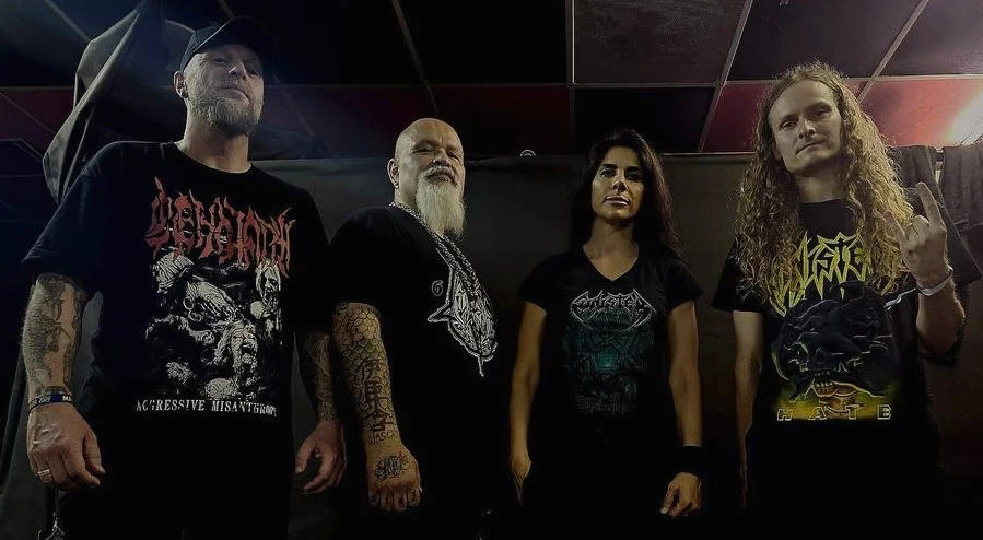 Sinister powraca do Polski Czterech ludzi pozuje frontalnie, ubrania czarnego koloru z widocznymi nadrukami zespołów metalowych na T-shirtach. Mężczyzna po lewej nosi czapkę z daszkiem, a mężczyzna po prawej ma długie, kręcone włosy. Tło jest ciemne i neutralne.