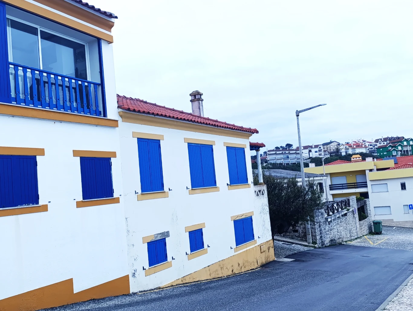 Budynek mieszkalny z niebieskimi okiennicami, usytuowany przy wąskiej ulicy. W tle widać inne domy na wzgórzu oraz częściowo zachmurzone niebo. Architektura typowa dla regionu śródziemnomorskiego, z białymi ścianami i czerwoną dachówką.