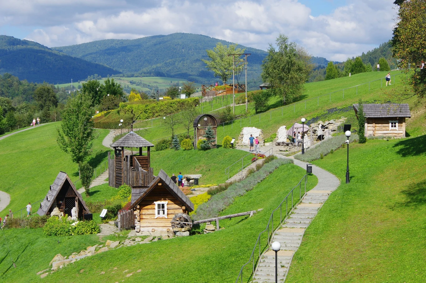 Drewniane domki na zielonym wzgórzu z widokiem na góry, otoczone kwiatami i drzewami, w tle majestatyczny górski krajobraz.