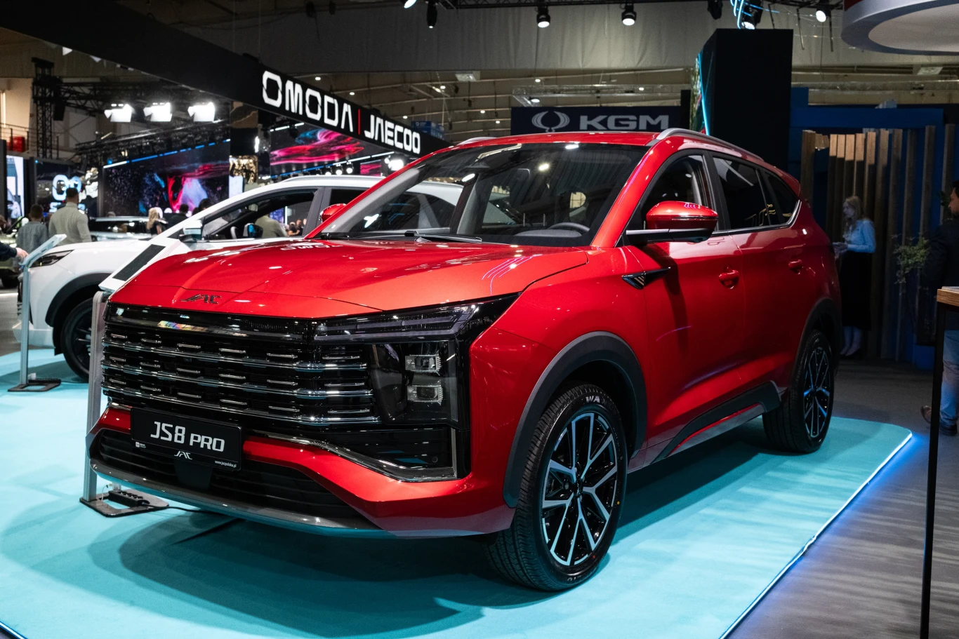 Czerwony samochód typu SUV model JS8 Pro stoi na wystawie targowej. Wokół niego widoczne są inne auta oraz logo znanej marki motoryzacyjnej. Samochód prezentuje się nowocześnie, z eleganckimi liniami i dużymi, czarnymi felgami.