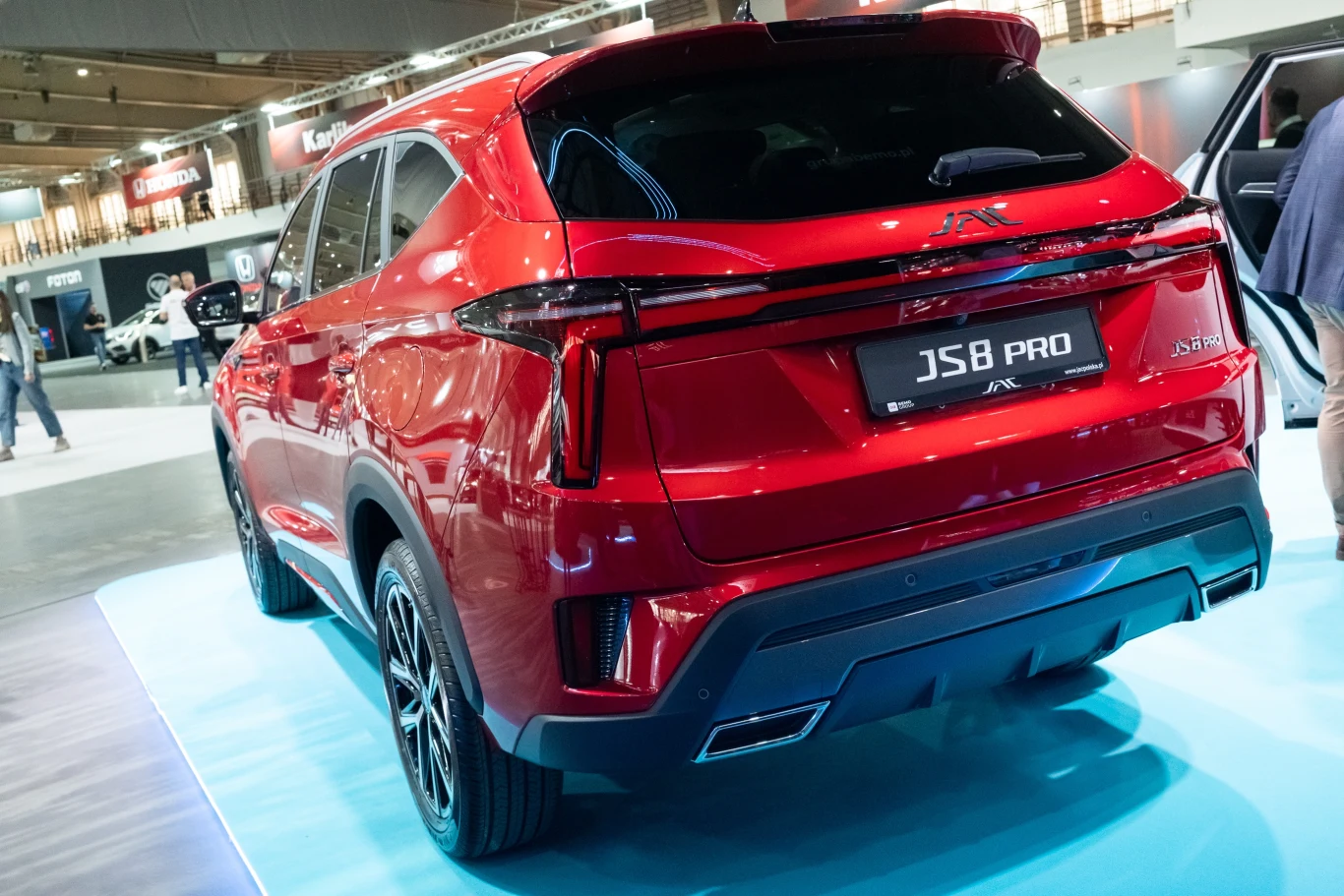 Czerwony samochód marki JS8 PRO zaprezentowany na wystawie motoryzacyjnej. Auto znajduje się na platformie, a w tle widoczne są inne pojazdy oraz wnętrze hali wystawowej.