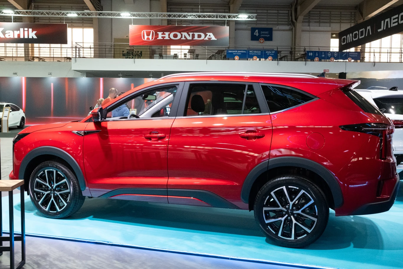 Czerwony SUV stoi na wystawie motoryzacyjnej, obok stołu. W tle widoczne są reklamy marek motoryzacyjnych, takich jak Honda i inne, oraz wnętrze hali wystawowej. Samochód ma nowoczesny i dynamiczny kształt.