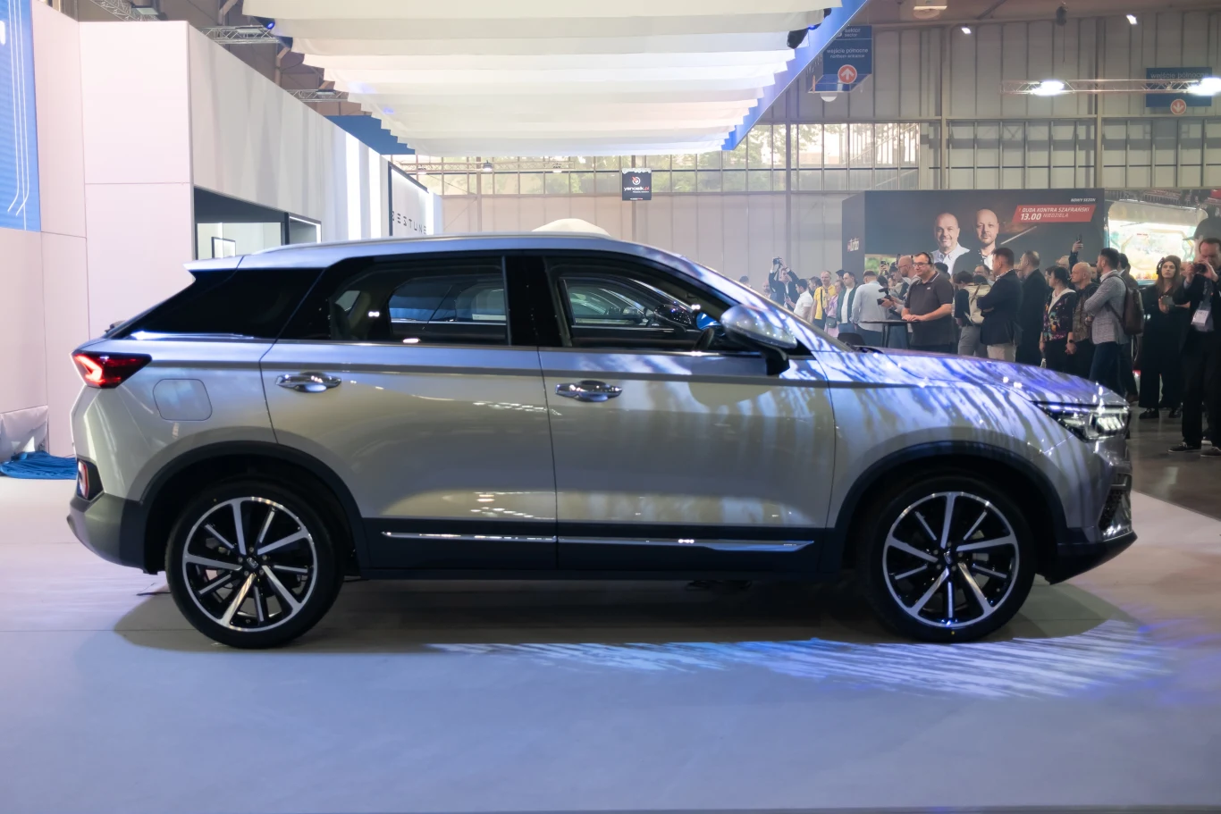 Szary SUV ustawiony bokiem na wystawie motoryzacyjnej, otoczony przez grupę ludzi oglądających pojazd i inne stanowiska.