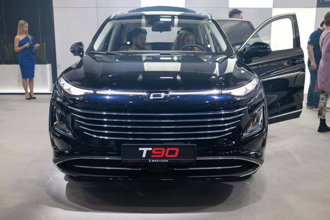 Czarny SUV T90 na tle białych ścian, z otwartymi przednimi drzwiami. Wewnątrz pojazdu siedzi mężczyzna i kobieta. W pobliżu stoi kilka osób w tle, z czego jedna osoba nosi niebieską sukienkę.