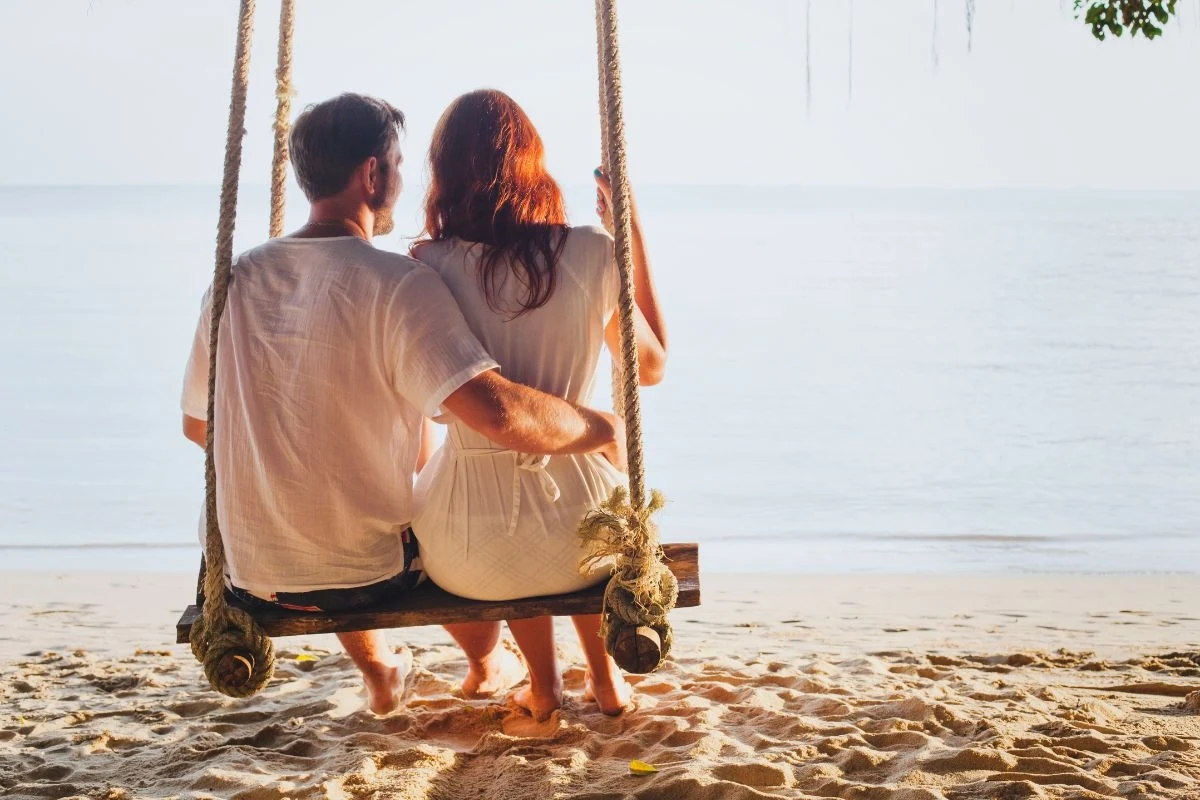 Para siedzi na huśtawce na plaży, spoglądając na morze. Kobieta ma długie rude włosy, oboje są ubrani na biało, co podkreśla spokojną i romantyczną atmosferę sceny. Przyjemne światło słoneczne tworzy ciepły nastrój.