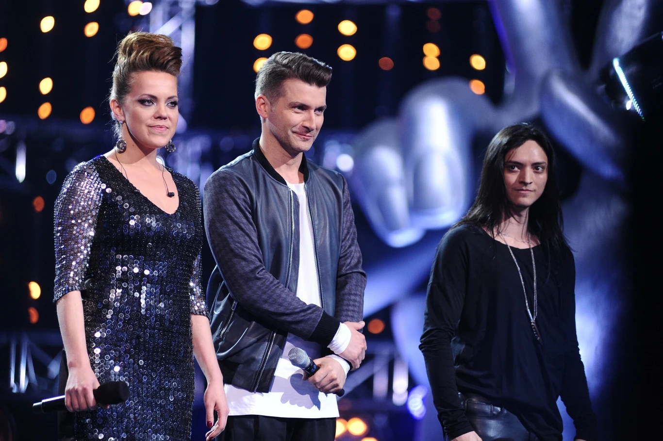 Trzy osoby stoją na scenie. Kobieta w eleganckiej sukience i dwaj mężczyźni w casualowych strojach. Publiczność w tle, atmosfera wydarzenia rozrywkowego. Gra świateł dodaje dynamiki, typowe dla programu muzycznego.