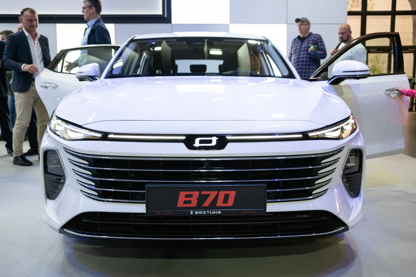 Biały samochód osobowy B70 marki Bestune na wystawie motoryzacyjnej, widoczny od frontu z otwartymi drzwiami. W tle kilku ludzi oglądających pojazd, skupiających się na jego detalach.