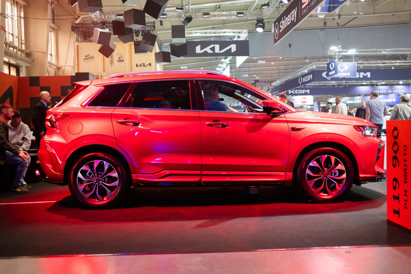 Czerwony SUV znajduje się na wystawie motoryzacyjnej, oświetlony intensywnym światłem. W tle widać ludzi oglądających pojazd oraz inne stoiska z nazwami różnych producentów samochodów. Pojazd stoi na podwyższonej platformie.