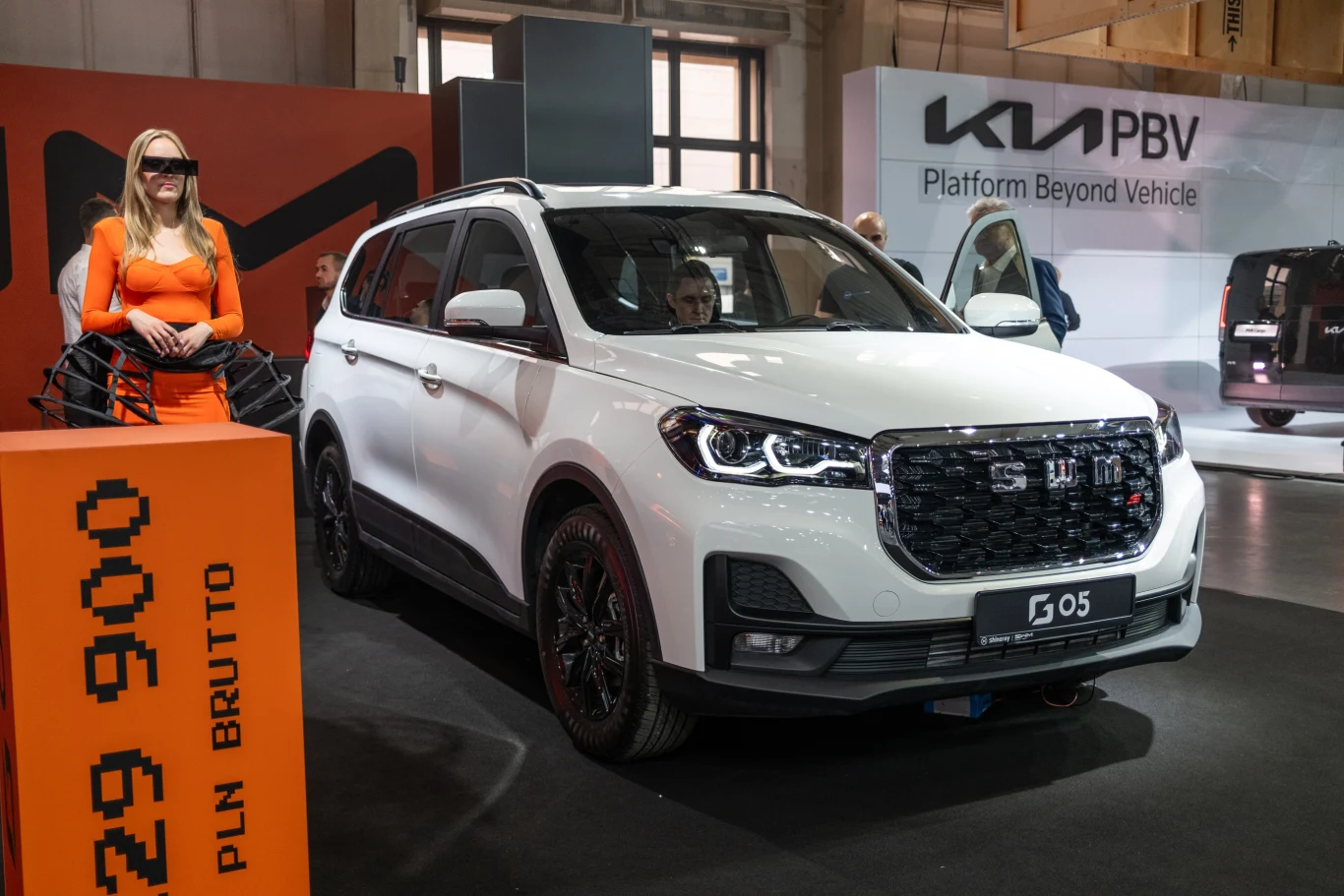 Stylowy, biały SUV na wystawie motoryzacyjnej, stojący obok kobiety w pomarańczowej sukience. Tło zawiera reklamy z nazwami marek.