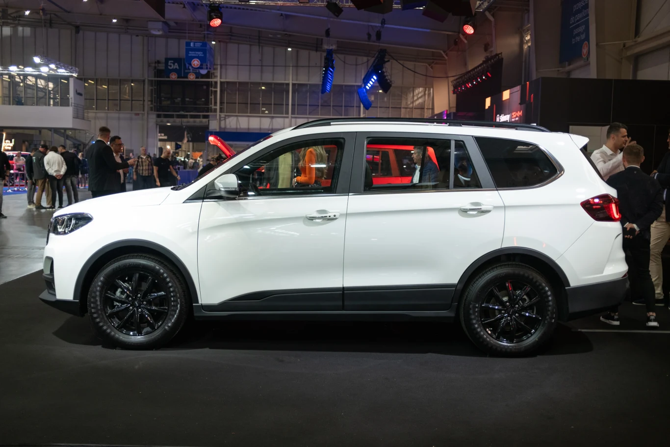 Biały SUV z czarnymi felgami na wystawie motoryzacyjnej. Pojazd stoi na platformie oświetlonej reflektorami w otoczeniu innych aut i osób zwiedzających. Tło stanowi hala wystawiennicza z widocznymi panelami świetlnymi i konstrukcjami stalowymi.