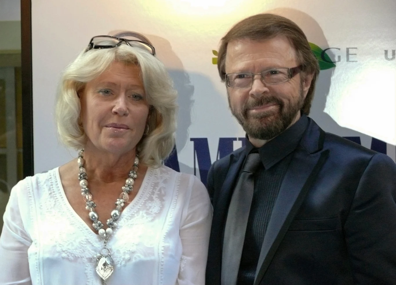 Przyjazne ujęcie dwóch uśmiechniętych osób stojących na tle napisu. Kobieta w eleganckiej białej bluzce z naszyjnikiem i okularami na głowie, mężczyzna w ciemnym garniturze i okularach. Obie postacie są wyraźne i dobrze oświetlone.