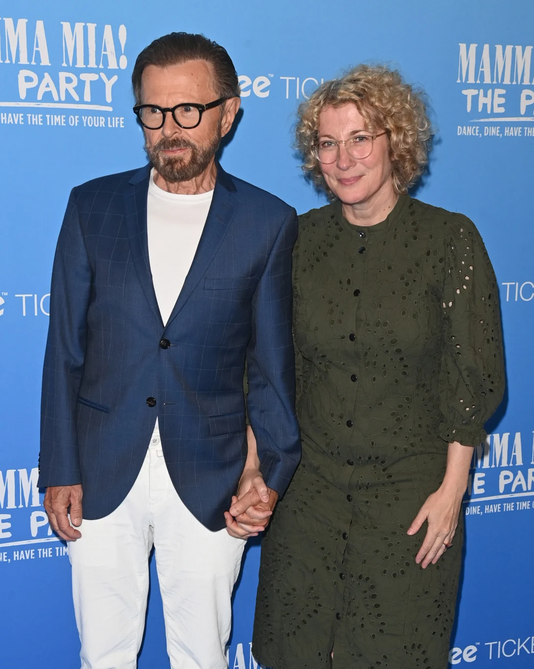 Mężczyzna i kobieta stoją razem na tle niebieskiej ściany z napisami związanymi z wydarzeniem Mamma Mia! Pomimo formalnych ubrań, atmosfera jest swobodna i radosna.