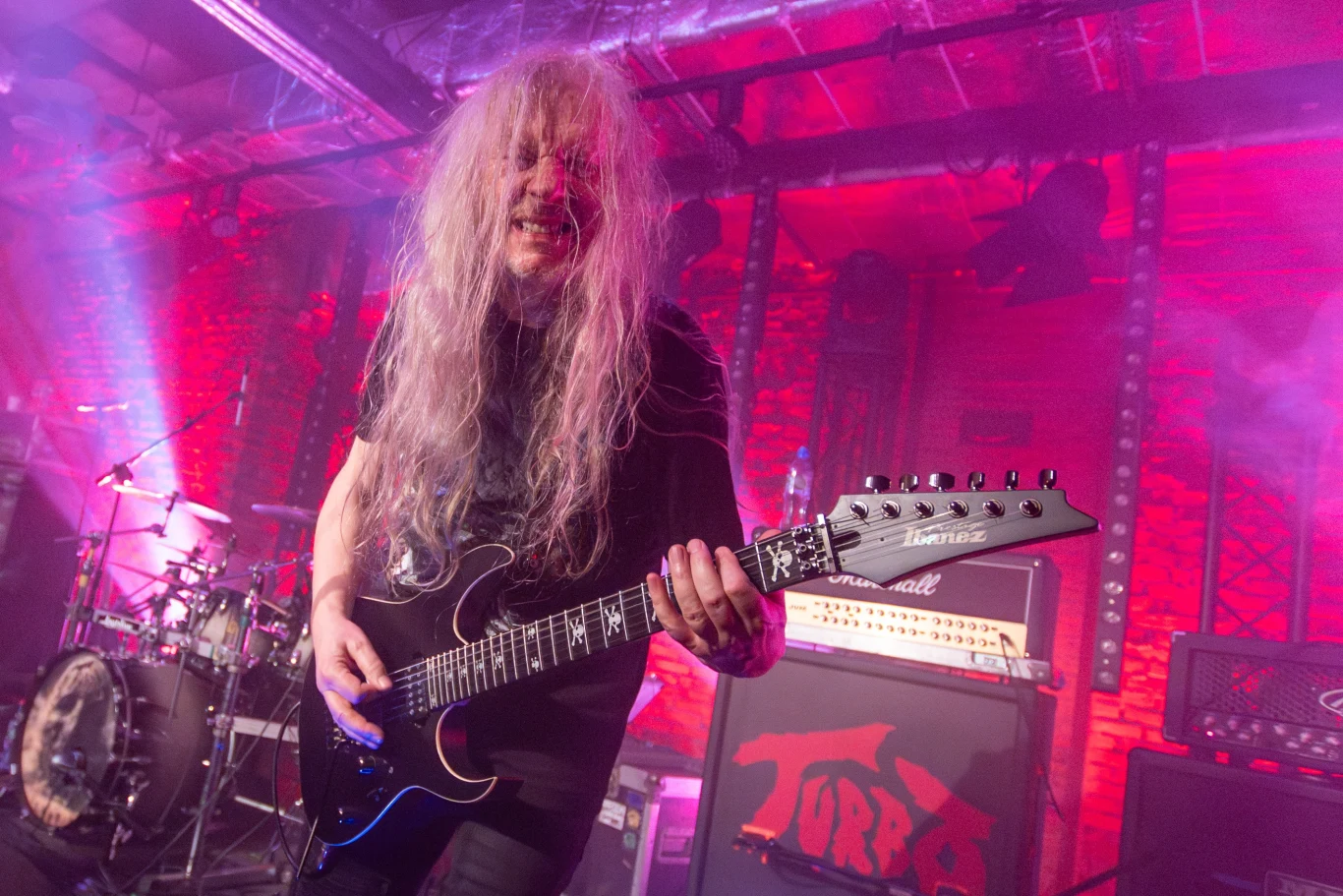 Muzyk z długimi, jasnymi włosami gra na gitarze elektrycznej na tle sceny oświetlonej czerwonymi światłami. Za nim widać perkusję i wzmacniacze gitarowe.