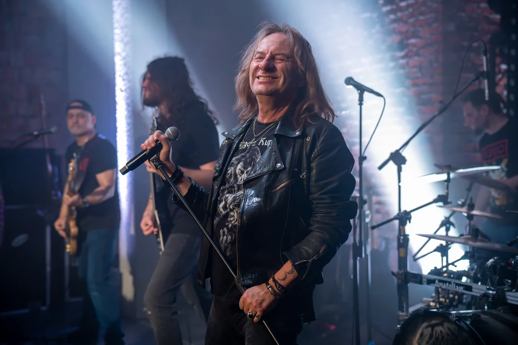 Grzegorz Kupczyk na gościnnych występach z Turbo, w tle od lewej Bogusz Rutkiewicz i Tomasz Struszczyk Grupa muzyków na scenie podczas koncertu rockowego. Frontman w skórzanej kurtce z mikrofonem w ręku, pozostali członkowie zespołu grają na instrumentach. Jasne światła sceniczne i ceglane ściany w tle tworzą rockowy klimat.