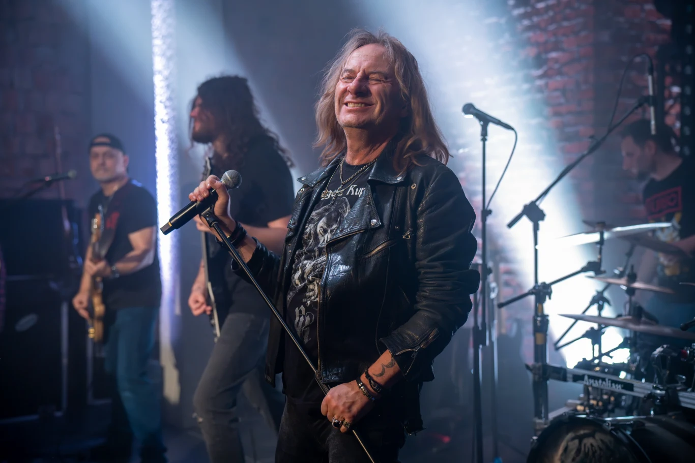 Grupa muzyków na scenie podczas koncertu rockowego. Frontman w skórzanej kurtce z mikrofonem w ręku, pozostali członkowie zespołu grają na instrumentach. Jasne światła sceniczne i ceglane ściany w tle tworzą rockowy klimat.