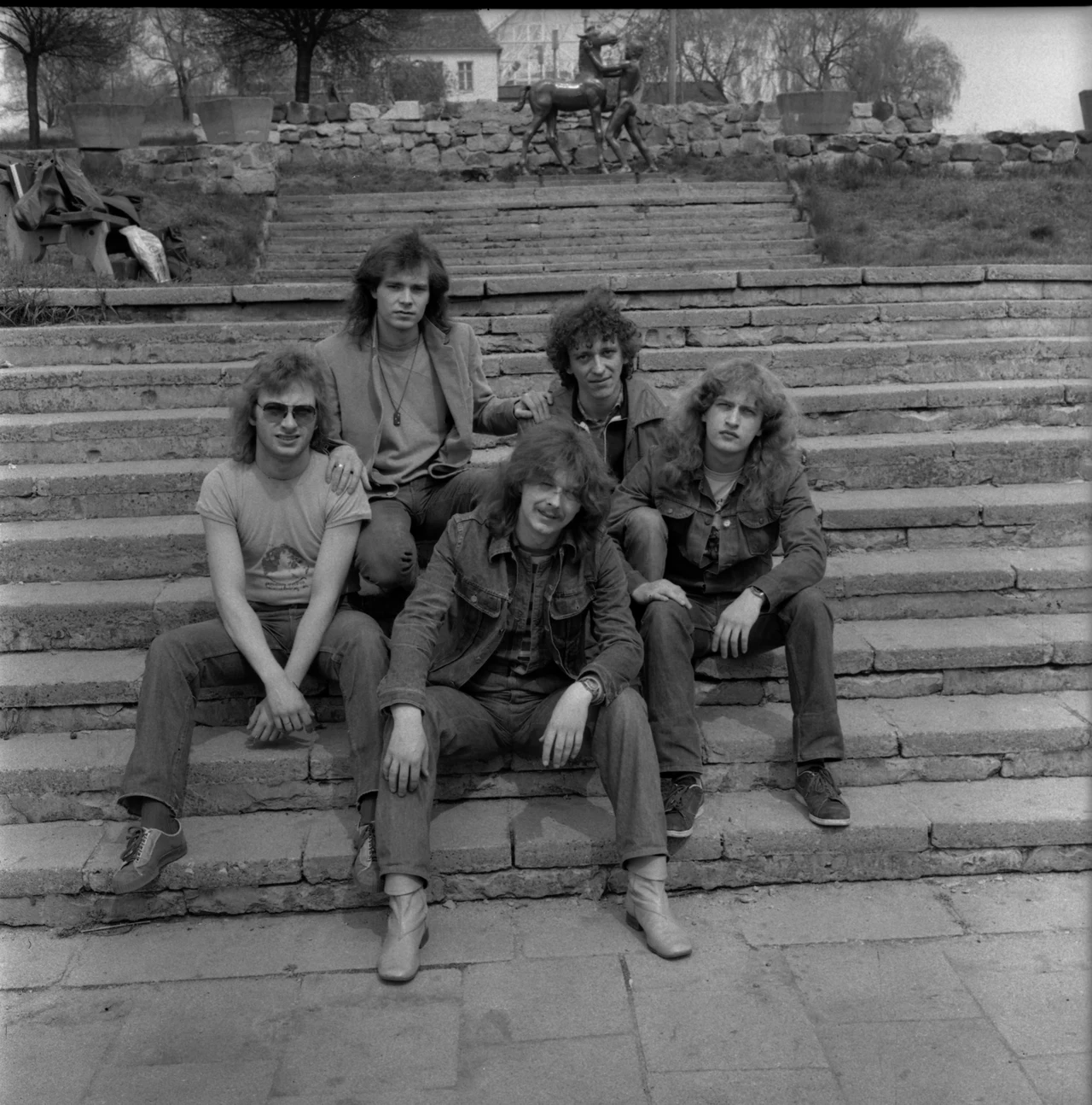 Turbo w połowie lat 80. - Wojciech Hoffmann w środku, pierwszy z lewej Grzegorz Kupczyk, pierwszy z prawej Bogusz Rutkiewicz Pięciu mężczyzn siedzi na schodach w plenerze, ubranych w dżinsy i kurtki, z bujnymi fryzurami z lat 70./80. W tle znajduje się rzeźba konia oraz zarośla. Atmosfera jest swobodna i nieformalna.