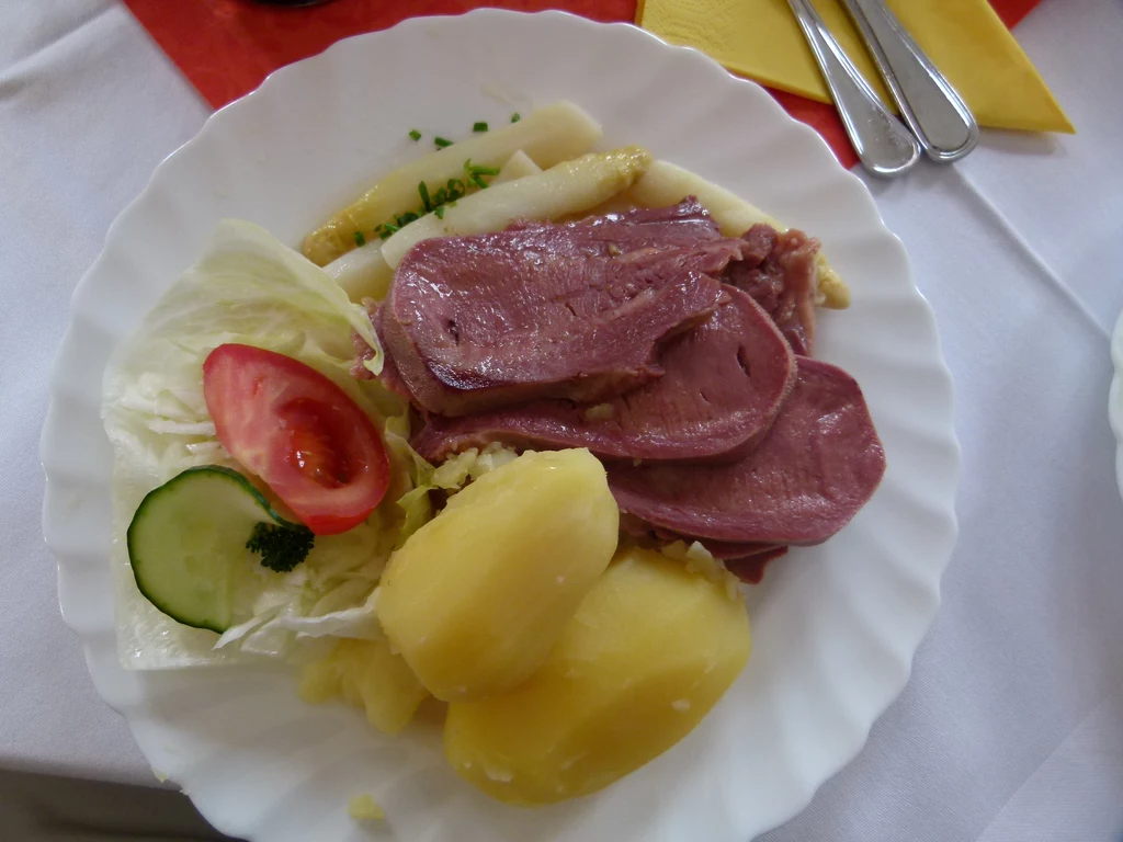 Danie z ozorem wołowym Danie składające się z plastrów wędzonego mięsa, ziemniaków, sałatki z pomidorem, ogórkiem. Obok białe szparagi. Wszystko na białym talerzu.