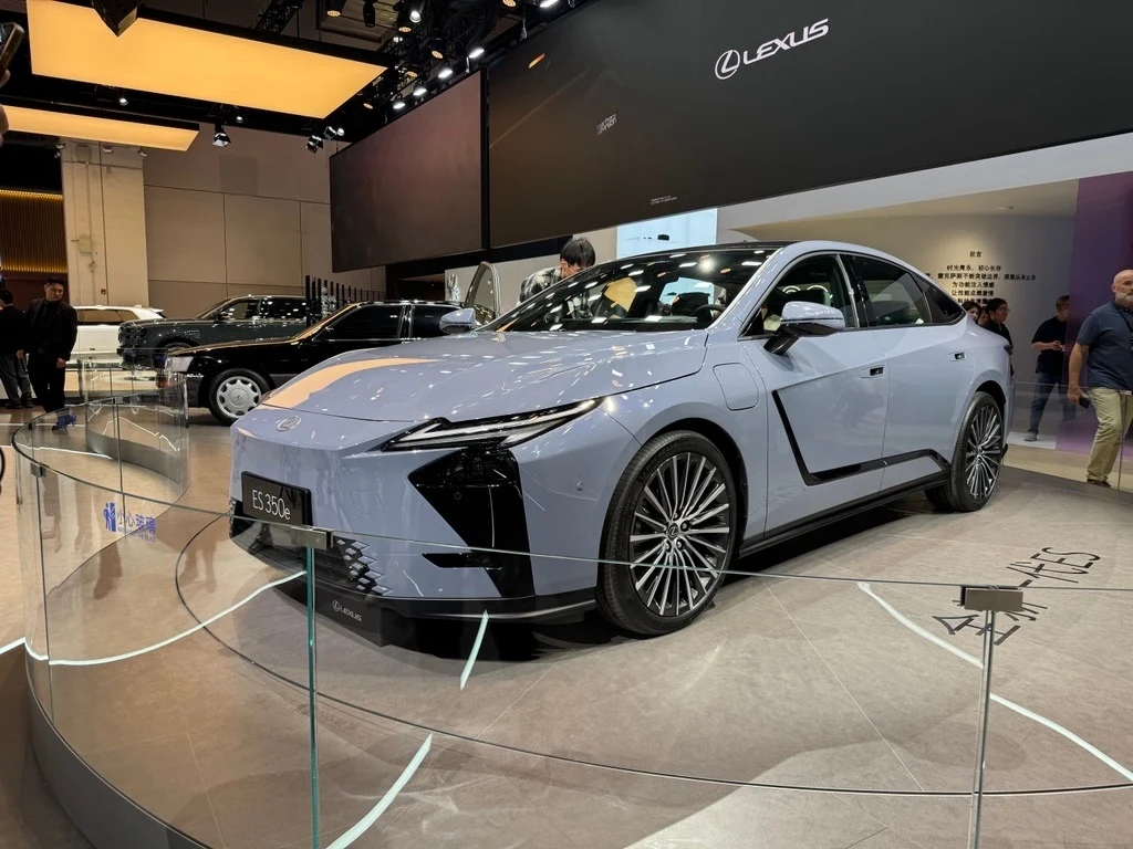 Luksusowy samochód marki Lexus, model RZ 350e w kolorze jasnoszarym, na wystawie motoryzacyjnej. Auto jest wyeksponowane na obrotowej platformie, wokół której znajdują się inni odwiedzający targi.