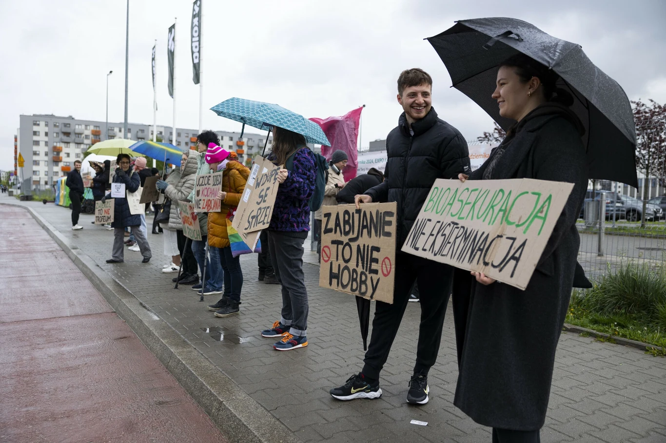 Grupa ludzi stoi na chodniku, trzymając transparenty z hasłami antyłowieckimi. Są ubrani w ciepłe ubrania i mają kolorowe parasole, co sugeruje, że jest deszczowy dzień. W tle widoczne są budynki mieszkalne.