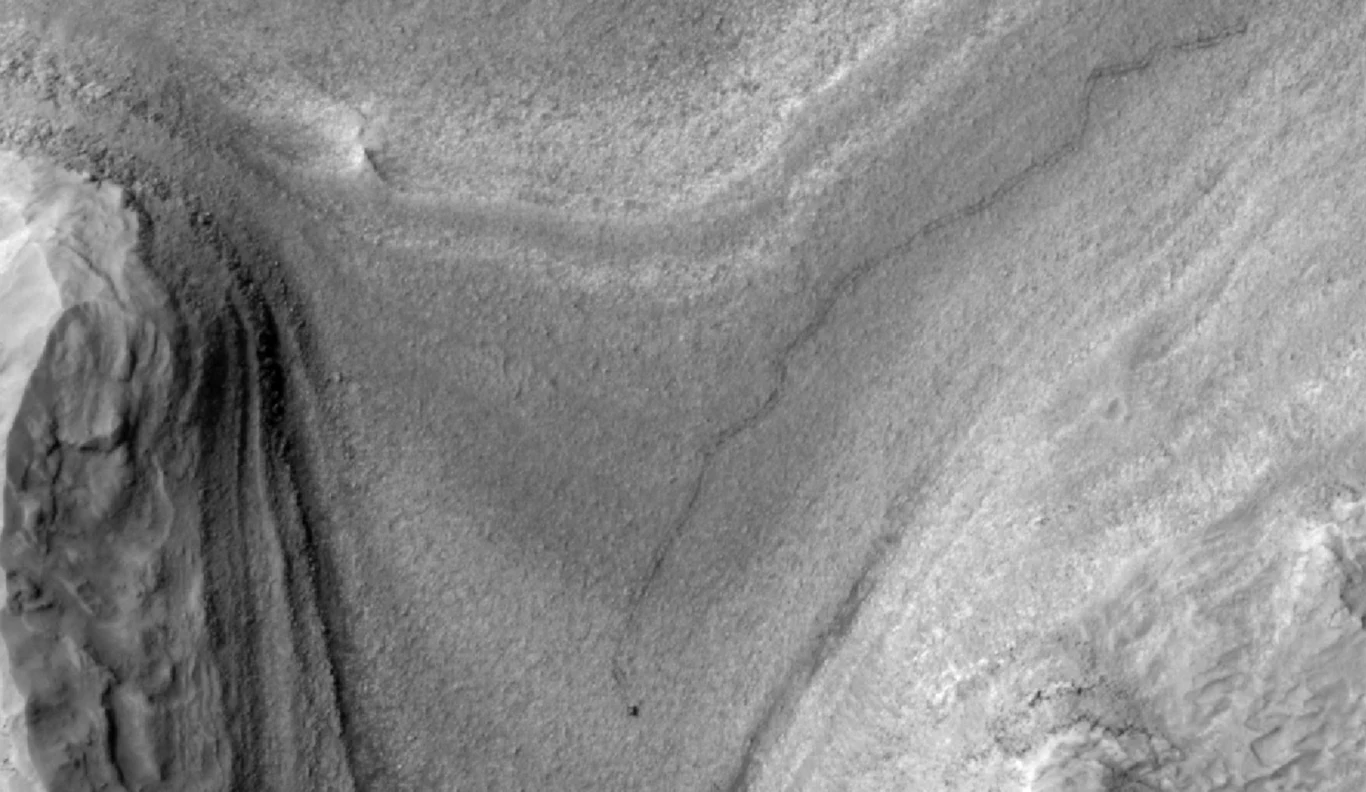 Łazik Curiosity i jego ślad na zdjęciu z Mars Reconnaissance Orbiter, które NASA uchwyciła pod koniec lutego. Formacje geologiczne o różnych warstwach i teksturach, widoczne w odcieniach szarości. Powierzchnia ma faliste kształty, które unoszą się i opadają, tworząc delikatne wzory przypominające linie konturowe na mapie.