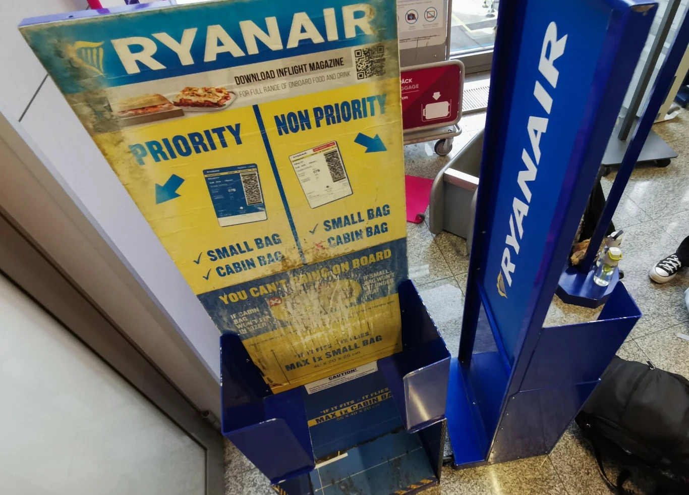 Stojak do mierzenia wymiarów bagażu podręcznego linii lotniczej Ryanair z wyraźnie oznaczonymi strefami dla bagażu priorytetowego i bez priorytetu, z instrukcjami dotyczącymi maksymalnych wymiarów torby i walizki.