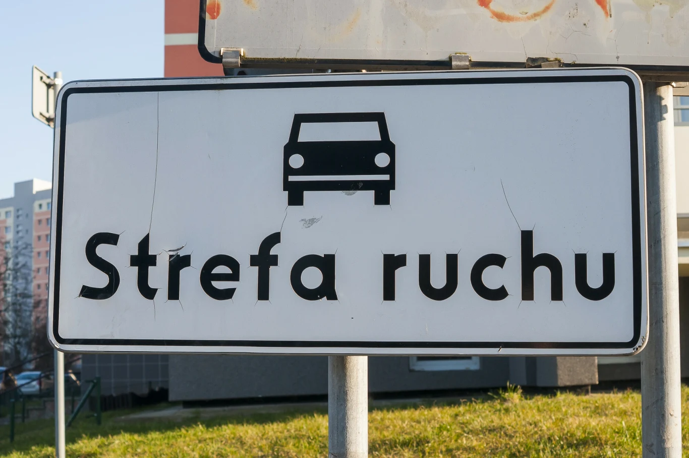 Znak drogowy informujący o strefie ruchu z ikoną samochodu na białym tle, umieszczony przy drodze w miejskim otoczeniu.