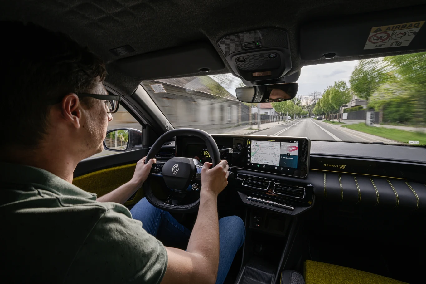 Mężczyzna prowadzi samochód Renault, korzystając z nowoczesnego systemu nawigacji widocznego na ekranie. Droga jest pusta, a krajobraz za oknem jest zielony i spokojny, co podkreśla komfort i technologię wnętrza auta.
