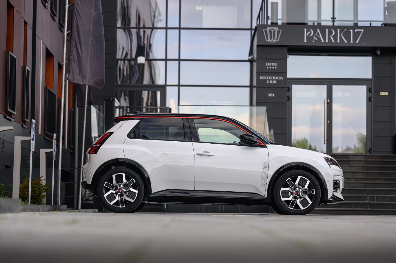 Biały samochód SUV zaparkowany przed nowoczesnym budynkiem z przeszklonymi drzwiami i napisami "Park17" oraz "Hotel, Restauracja, Bar". Auto znajduje się na szerokim chodniku, wokół widać elementy miejskiej architektury.