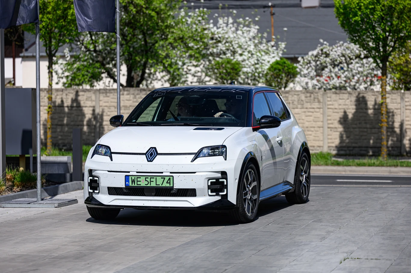 Biały samochód marki Renault o sportowym wyglądzie, stojący na ulicy w otoczeniu zielonych drzew i kwitnących krzewów. Tablica rejestracyjna wskazuje lokalizację w Polsce.