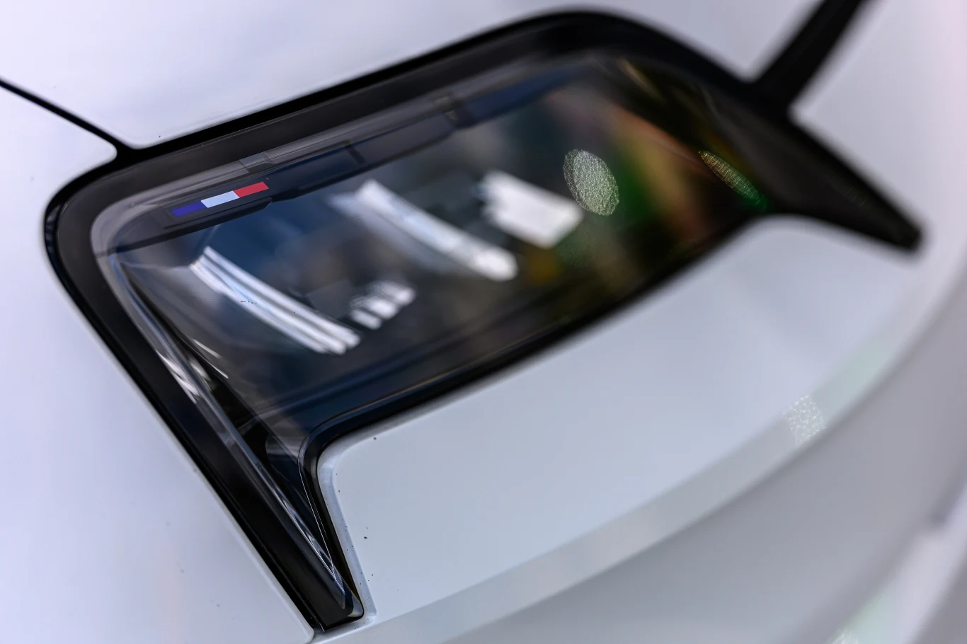 zbliżenie na reflektor samochodowy z nowoczesnym, minimalistycznym designem, umieszczony na jasnym tle karoserii. Detale reflektora są wyraźne i ukazują jego precyzyjną konstrukcję, włącznie z charakterystycznymi liniami i załamaniami światła.